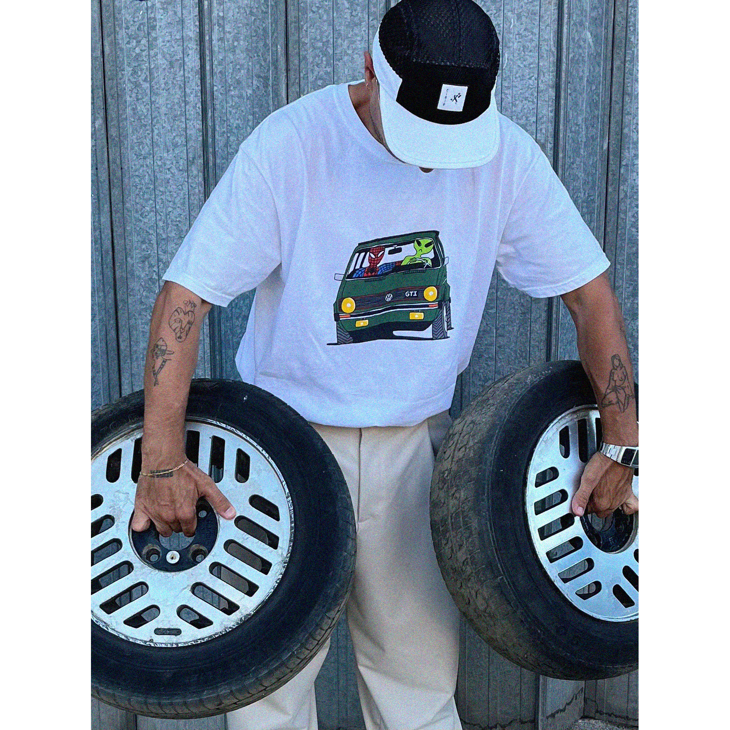 Model wearing the Unisex Ring-Spun Cotton Crewneck Heavyweight T-Shirt in white, depicting Spiderman and a green alien on a road trip in a vintage green Volkswagen Golf, holding two car wheels